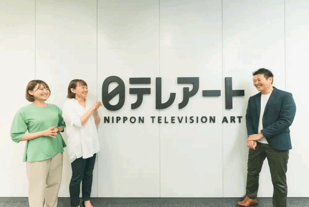 株式会社日本テレビアート様の支援事例の写真。日テレアートのロゴが掲げられた壁の前に担当者3名が並んでいる